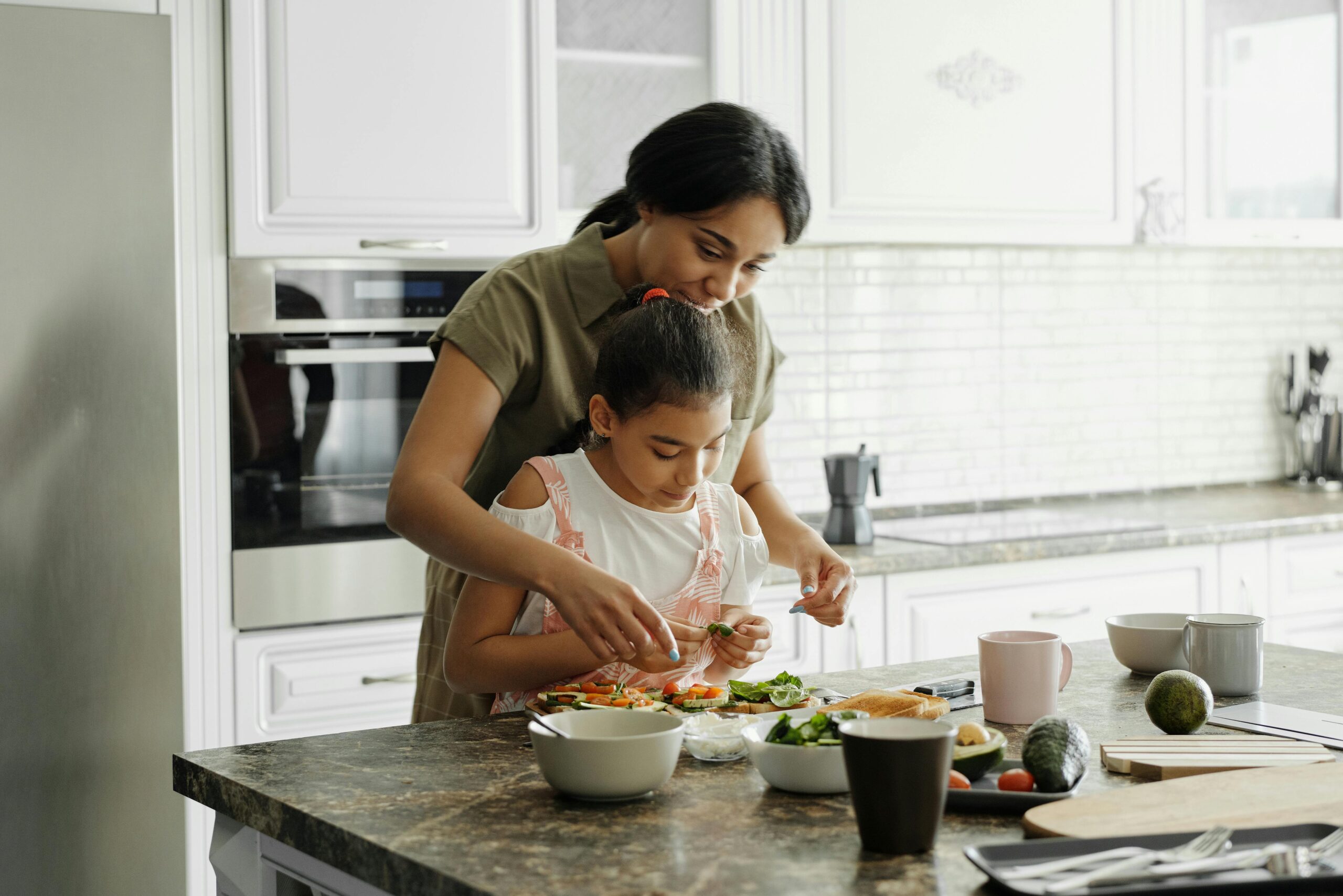 A dark haired woman and a child make a smoothie together in a clean, white kitchen